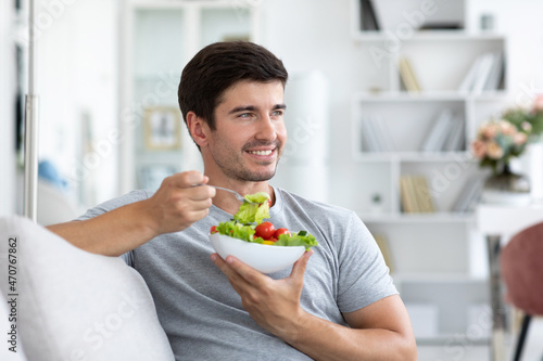 Handsome man eating fresh vegetable salad while sitting on the couch at home, healthy and vegan