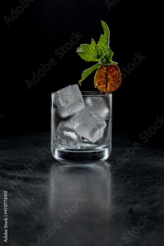 A rock glass with ice on a black background in the backlight. Decorated with a skewer with orange chip and mint.