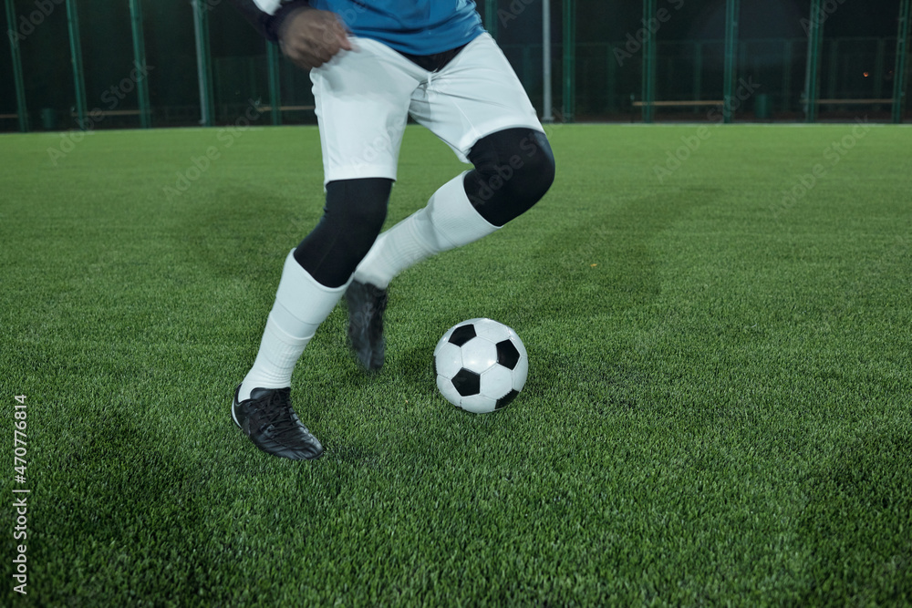 Legs of young active football player running after soccer ball while playing or training on green lawn at stadium