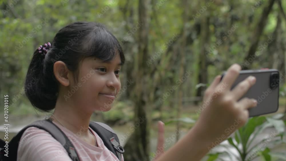 Asian cute girl with backpack enjoy taking a selfie with smartphone while posing near a creek in tropical forest during summer. Outdoor nature camping. Jungle adventure. Young lifestyle. Thailand.
