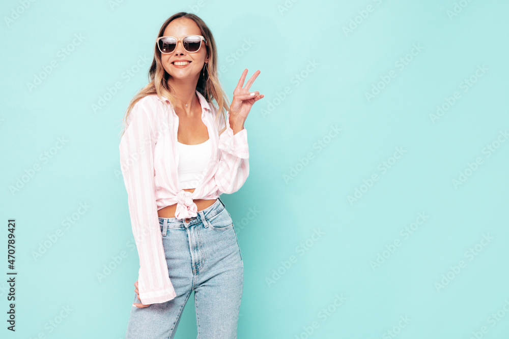 Young beautiful smiling female in trendy summer clothes. Sexy carefree woman posing near blue wall in studio. Positive model having fun indoors. Cheerful and happy. Shows peace sign, in sunglasses Young beautiful smiling female in trendy summer clothes. Sexy carefree woman posing near blue wall in studio. Positive model having fun indoors. Cheerful and happy. Shows peace sign, in sunglasses
