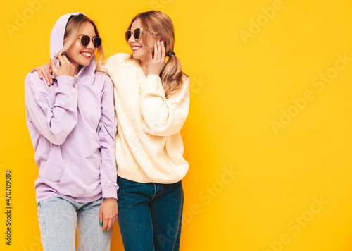 Two young beautiful smiling blond hipster female in trendy summer clothes. Sexy carefree women posing near yellow wall in studio. Positive models having fun. Cheerful and happy. In sunglasses