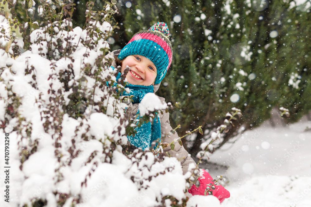 winter walking, children winter holiday. happy smiling girl child playing in snow in winter park, wearing a bright knitted hat and scarf and waterproof mittens. winter outdoor concept