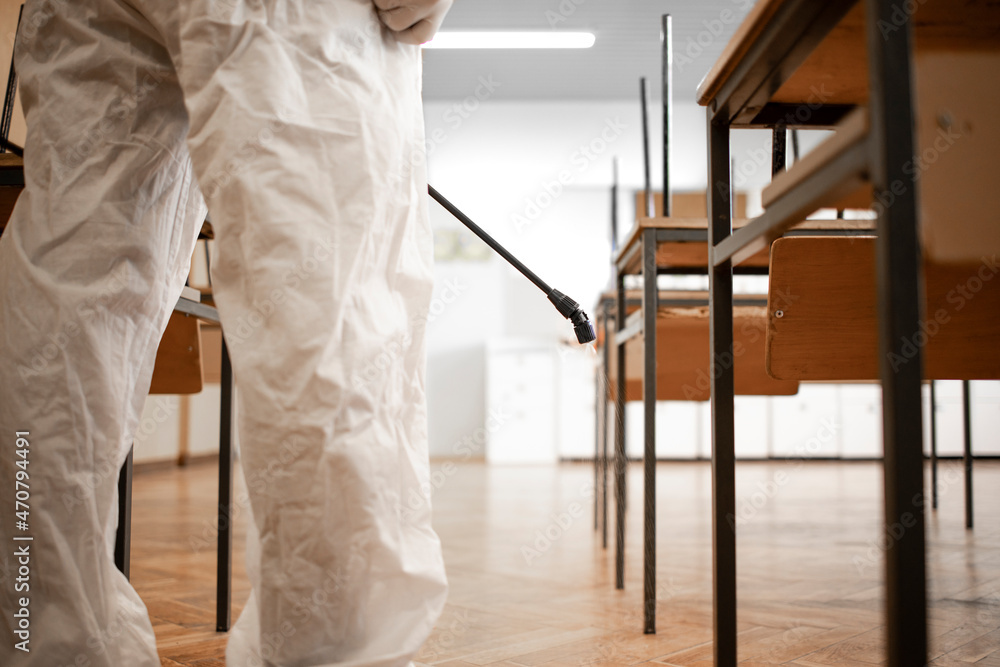 Worker in white sterile protection suit disinfecting and sanitizing desks and chairs in school classroom during corona virus pandemic.