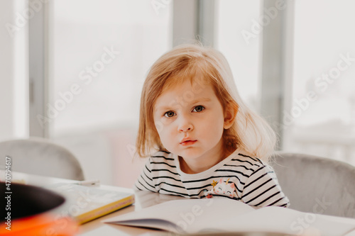 Cute toddler girl sitting in kitchen and drawing while her mother cooking.
