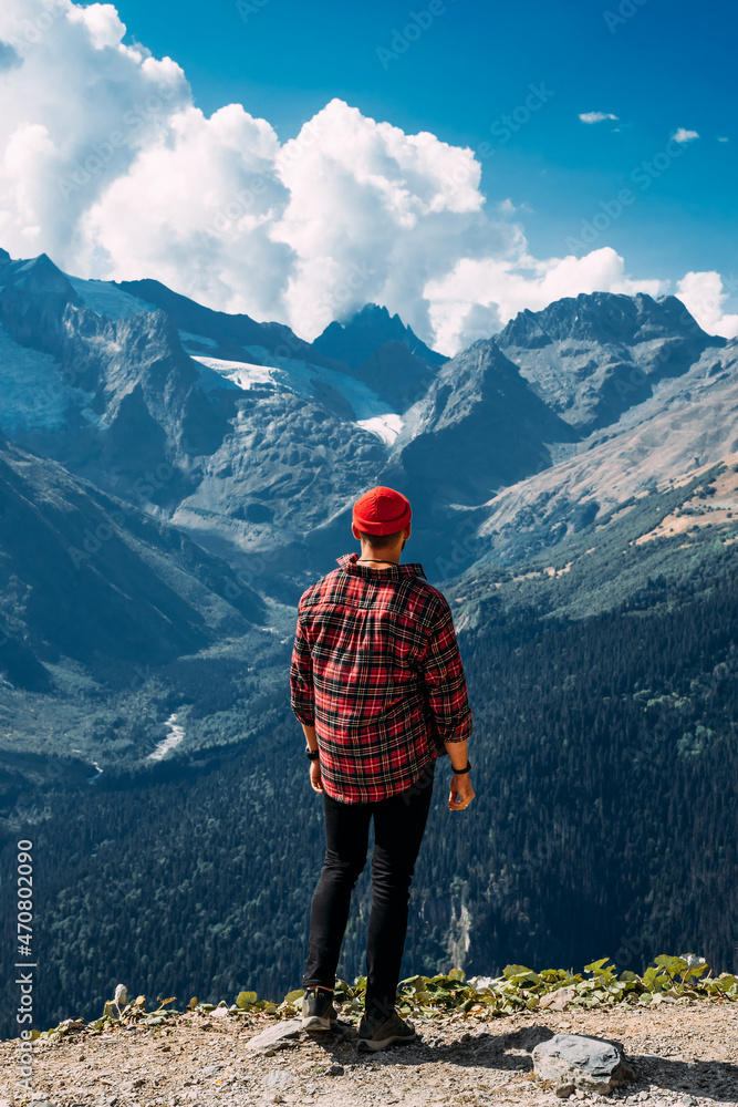 A traveler on the background of mountains. A tourist stands on top of a mountain. Male traveler in the mountains, rear view. A traveler in a red plaid shirt and a red hat. Domestic tourism. Copy space