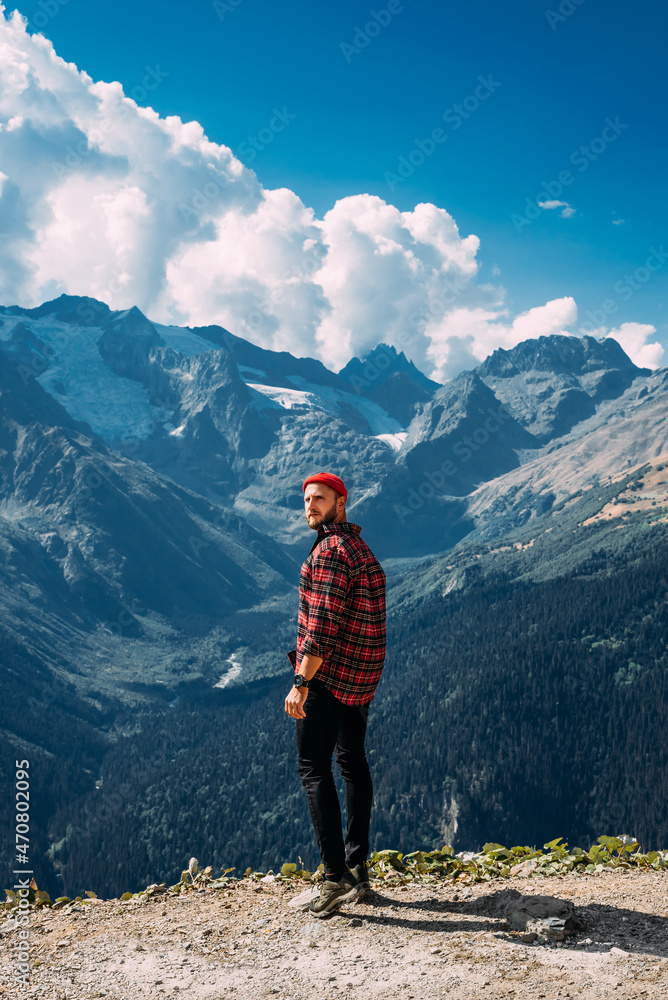 Stylish bearded man on top of a mountain. Portrait of a traveler in a red cap and a plaid shirt against the background of mountains. The concept of recreation and tourism. Travel. Copy space