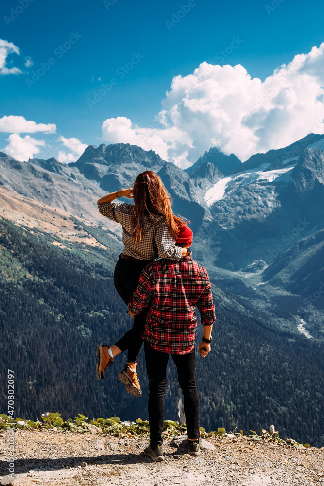 A traveling couple in plaid shirts against the backdrop of mountains. Two tourists on the top of the mountain. A man and a woman in the mountains. Two travelers enjoy climbing the mountain. Copy space A traveling couple in plaid shirts against the backdrop of mountains. Two tourists on the top of the mountain. A man and a woman in the mountains. Two travelers enjoy climbing the mountain. Copy space