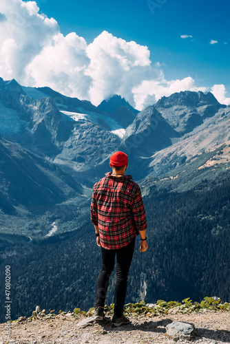 A traveler on the background of mountains. A tourist stands on top of a mountain. Male traveler in the mountains, rear view. A traveler in a red plaid shirt and a red hat. Domestic tourism. Copy space