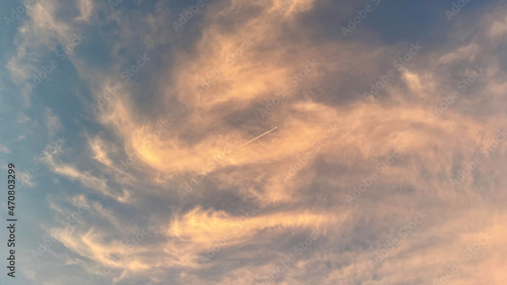 Plane flies high in the sky, leaving a long white trail behind it. Heaven and sunset. Beautiful clouds. Colored glow of the sun. Airplane in skies. Twilight at the evening. Gray orange yellow colours.