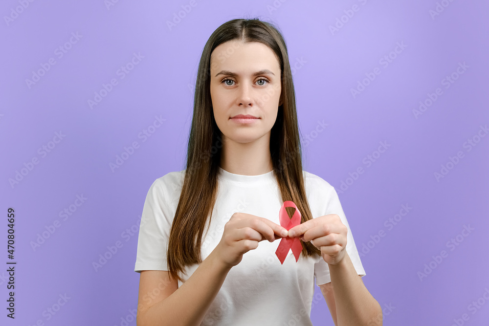 Kind cute young woman holding pink ribbon, symbol of breast cancer awareness, recommendation and warning of cancer prevention, female health diagnostics, isolated over violet background in studio Kind cute young woman holding pink ribbon, symbol of breast cancer awareness, recommendation and warning of cancer prevention, female health diagnostics, isolated over violet background in studio