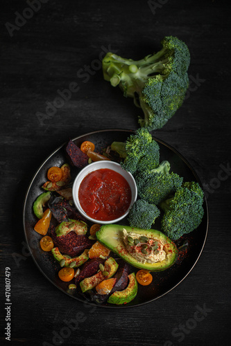 Vegan snack plate with broccoli, baked beet, orange, cherry tomatoes, avocado, hummus and tomato sauce. Black wooden background. Top view.