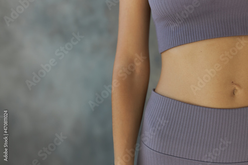 Crop shot of female belly button and hand, dressed in violet short top and leggings, isolated over grey textured wall with copy space for your advertising content. Perfect human body photo