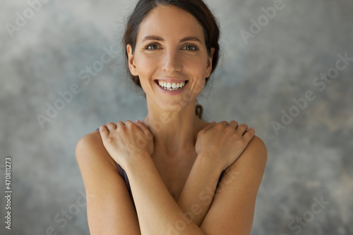 Portrait of joyful, happy elegant, enigmatic female of 30s standing with arms on her shoulders covering naked upper side of body with crossed hands, smiling cheerfully, having light make-up
