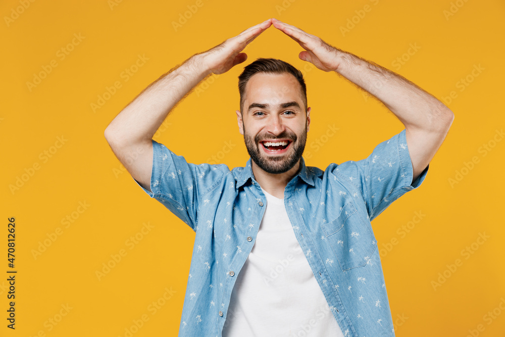 Young smiling happy fun man 20s wearing blue shirt white t-shirt hold folded hands above head like house roof, stay home isolated on plain yellow background studio portrait. People lifestyle concept. Young smiling happy fun man 20s wearing blue shirt white t-shirt hold folded hands above head like house roof, stay home isolated on plain yellow background studio portrait. People lifestyle concept.