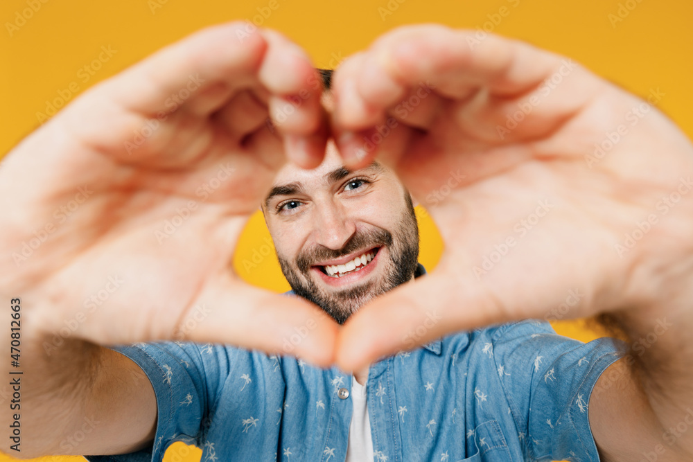 Close up romantic fun young smiling happy man 20s in blue shirt showing shape heart look through hands heart-shape sign isolated on plain yellow background studio portrait. People lifestyle concept. Close up romantic fun young smiling happy man 20s in blue shirt showing shape heart look through hands heart-shape sign isolated on plain yellow background studio portrait. People lifestyle concept.
