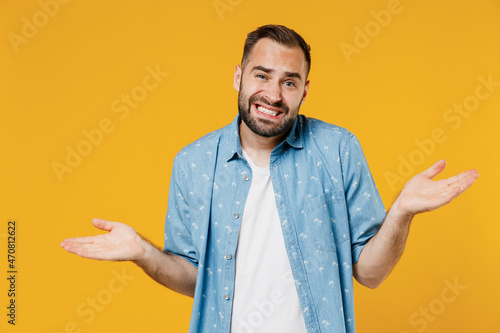 Young confused sad man 20s wearing blue shirt shrugging shoulders looking puzzled, have no idea, nothing to say, standing questioned and unaware isolated on plain yellow background studio portrait