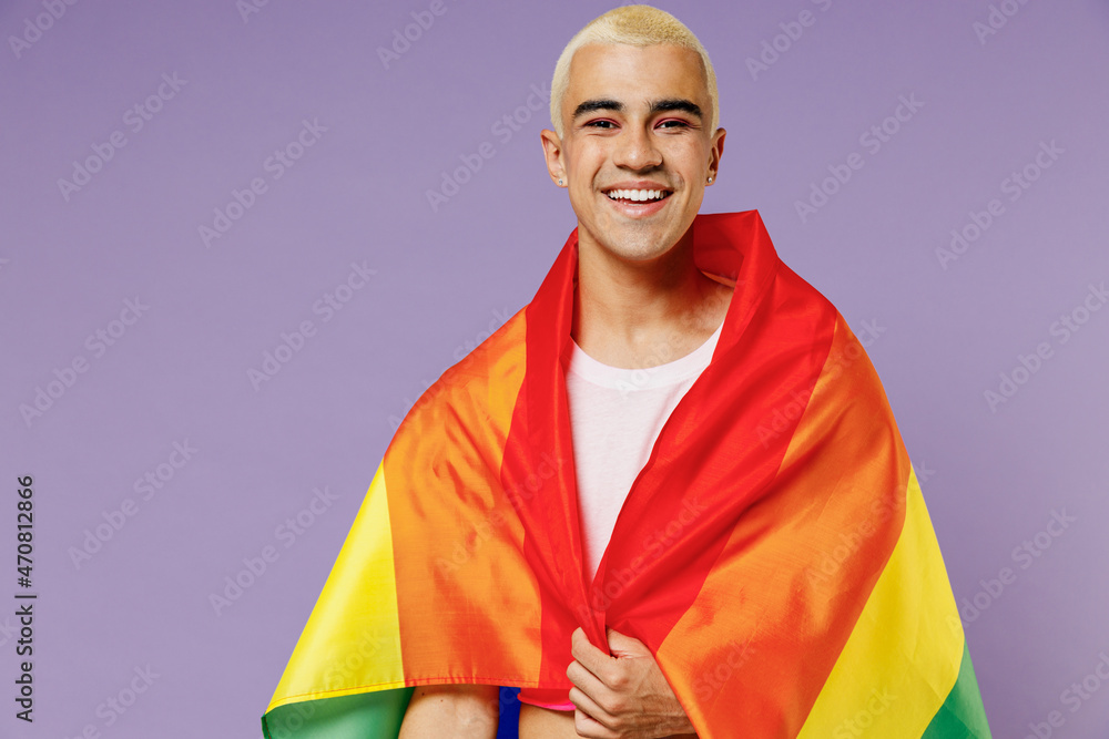 Young smiling hispanic latin gay man 20s with make up wrapped in rainbow striped flag look camera isolated on plain pastel purple background studio portrait. People lifestyle fashion lgbtq concept