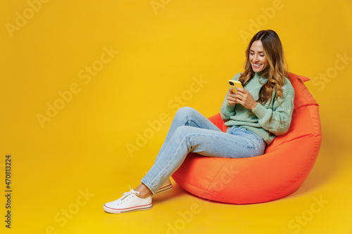 Full body young smiling woman 30s in green knitted sweater sit in bag chair hold use mobile cell phone chatting browsing internet isolated on plain yellow background studio. People lifestyle concept.