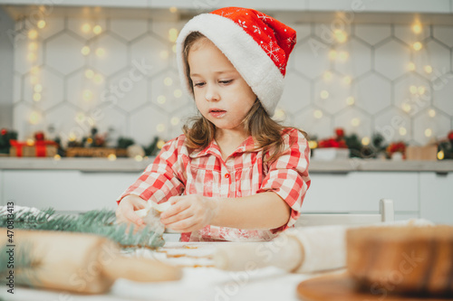 Dark-haired girl 3 years old in red Christmas cap and checkered shirt cuts out gingerbread cookies from rolled dough
