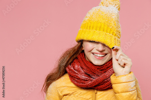 Smiling fascinating fun young woman 20s years old wears yellow jacket hat mittens pulling cap with hand cover close hiding eyes with hat isolated on plain pastel light pink background studio portrait.