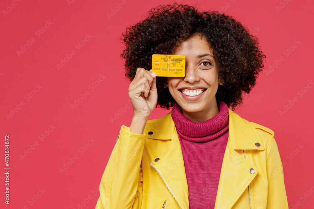 Satisfied young curly black latin woman 20s wears yellow jacket hold in hand close cover eye with credit bank card isolated on plain red background studio portrait. Achievement career wealth concept. Satisfied young curly black latin woman 20s wears yellow jacket hold in hand close cover eye with credit bank card isolated on plain red background studio portrait. Achievement career wealth concept.