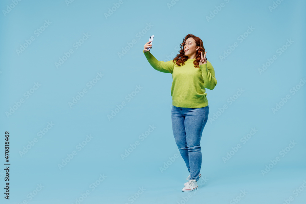 Full body smiling young chubby overweight plus size big fat fit woman wear green sweater doing selfie shot on mobile cell phone show v-sign isolated on plain blue background People lifestyle concept. Full body smiling young chubby overweight plus size big fat fit woman wear green sweater doing selfie shot on mobile cell phone show v-sign isolated on plain blue background People lifestyle concept.