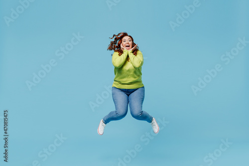 Fototapeta Naklejka Na Ścianę i Meble -  Young overjoyed fun excited surprised chubby overweight plus size big fat fit woman wear green sweater jump high hold face isolated on plain blue background studio portrait. People lifestyle concept