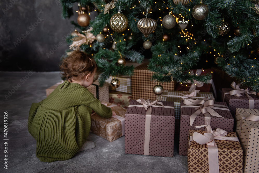 Little girl in green warm dress opens boxes of gifts in craft paper under a Christmas tree.