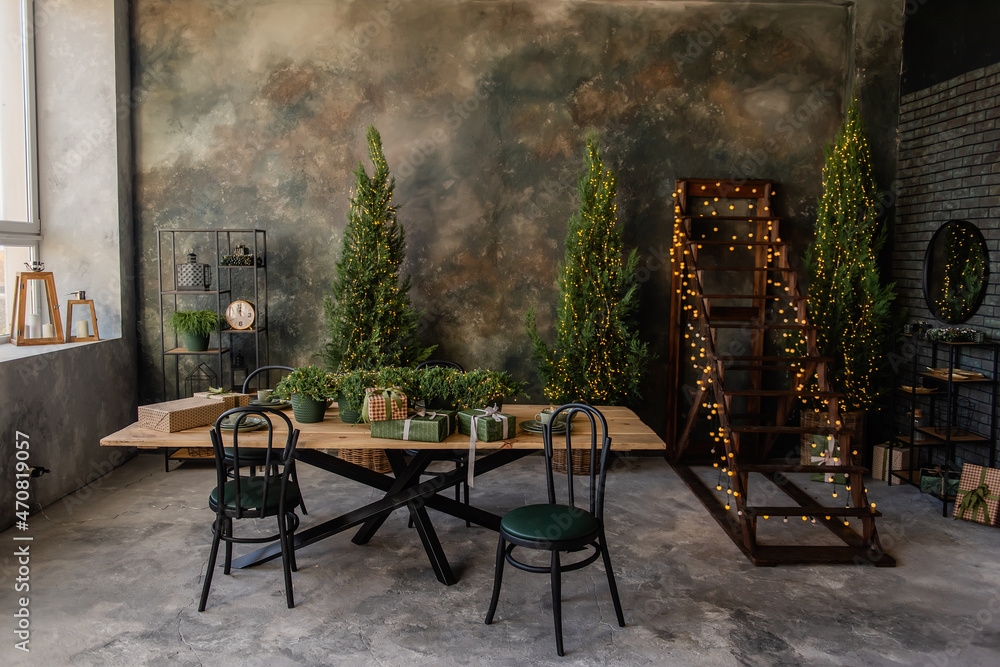 The dark christmas loft has wooden table with gifts, black chairs, stylish clock rack. Grunge wall in green-gray color. Trendy modern interior, ladder with garlands. Business cabinet. Copy space