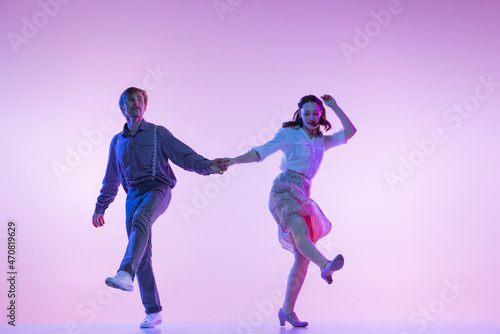 Dynamic portrait of young dancers, man and woman in vintage style outfits dancing swing isolated on lilac color background in neon light