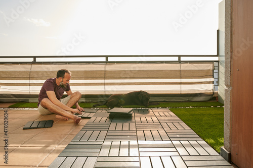middle-aged man setting up an artificial floor on his terrace with his Labrador retriever laying next to him