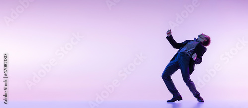 Portrait of young stylish man in vintage retro style clothes dancing swing dance isolated on lilac color background in neon light