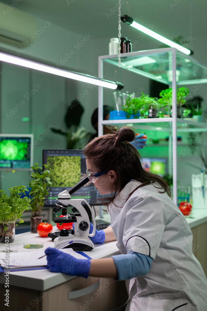 Biologist researcher doctor analyzing gmo plants sample using medical microscope writing biological expertise on clipboard. Botanist scientist woman working in biochemistry hospital laboratory Biologist researcher doctor analyzing gmo plants sample using medical microscope writing biological expertise on clipboard. Botanist scientist woman working in biochemistry hospital laboratory