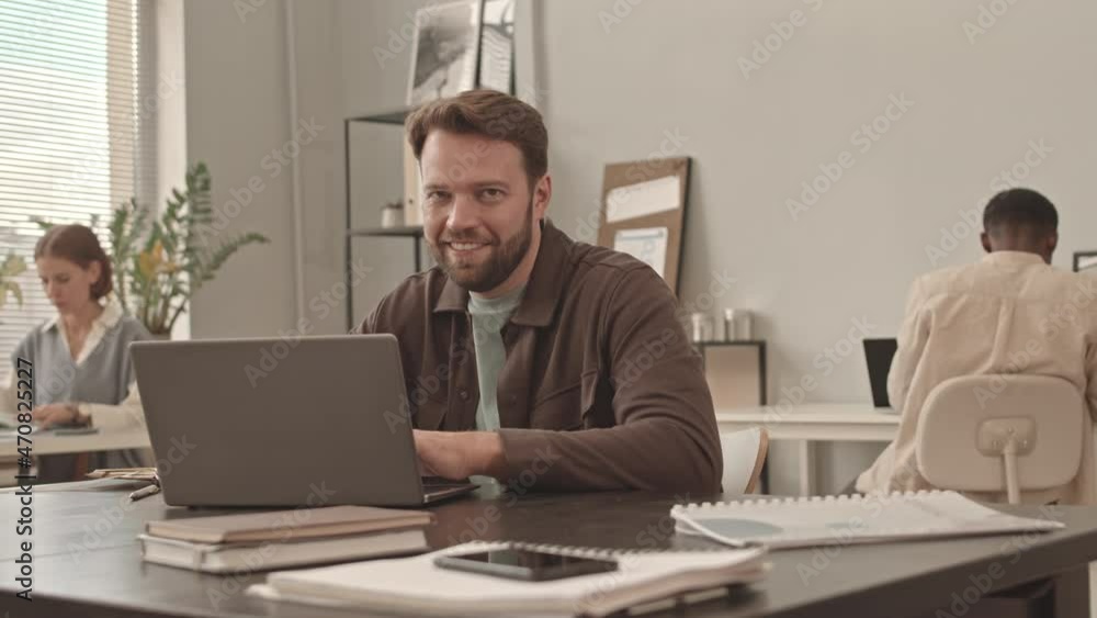 Medium close-up of young bearded Caucasian male manager sitting at desk in office, using laptop computer, then looking and smiling on camera Medium close-up of young bearded Caucasian male manager sitting at desk in office, using laptop computer, then looking and smiling on camera