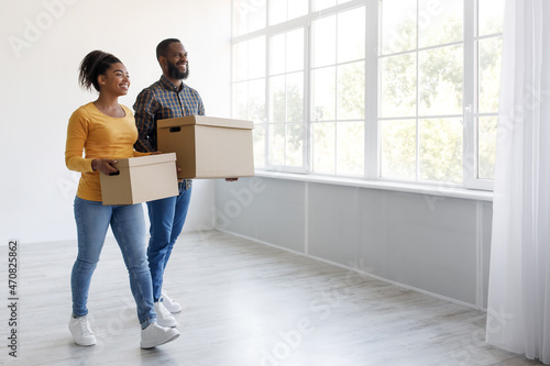 Cheerful glad millennial african american husband and wife carry cardboard boxes and moving