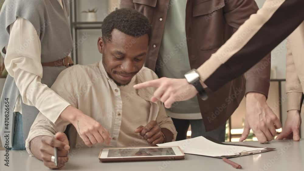 Waist-up of young male African American designer sitting at desk in office at daytime, three cropped coworkers standing behind him and pointing fingers at tablet computer Waist-up of young male African American designer sitting at desk in office at daytime, three cropped coworkers standing behind him and pointing fingers at tablet computer