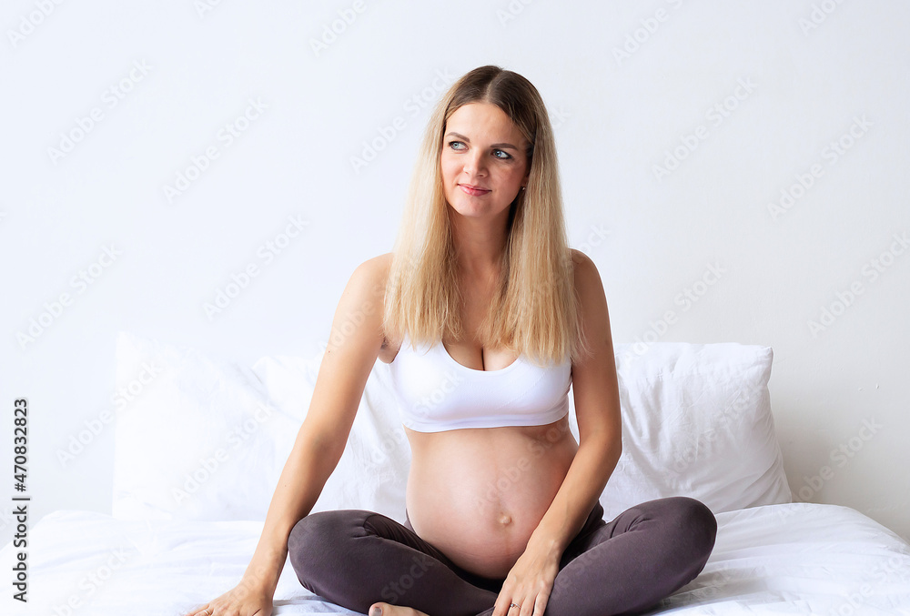Cute blonde pregnant girl sitting on the bed at home. On a light background. Portrait. 8 month of pregnancy. The concept of waiting for the birth of a baby. Copyspace. Cute blonde pregnant girl sitting on the bed at home. On a light background. Portrait. 8 month of pregnancy. The concept of waiting for the birth of a baby. Copyspace.