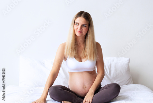 Cute blonde pregnant girl sitting on the bed at home. On a light background. Portrait. 8 month of pregnancy. The concept of waiting for the birth of a baby. Copyspace.