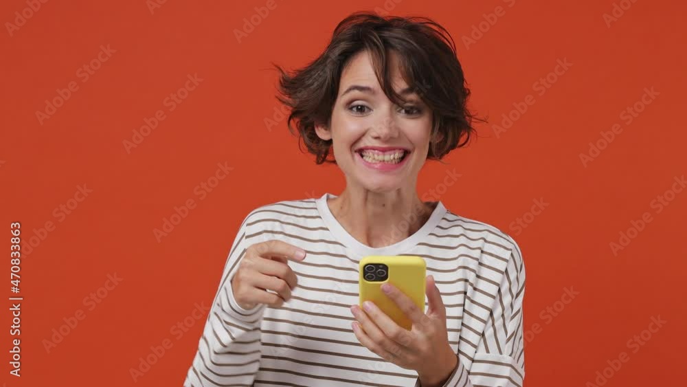 Happy surprised fun young brunette woman 20s years old wears striped shirt hold use mobile cell phone pointing finger on blank screen workspace area isolated on plain orange background studio portrait Happy surprised fun young brunette woman 20s years old wears striped shirt hold use mobile cell phone pointing finger on blank screen workspace area isolated on plain orange background studio portrait