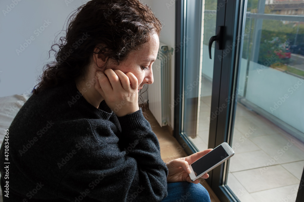 Side view of a sad woman checking phone sitting on the bed in the bedroom at home next to a window Side view of a sad woman checking phone sitting on the bed in the bedroom at home next to a window