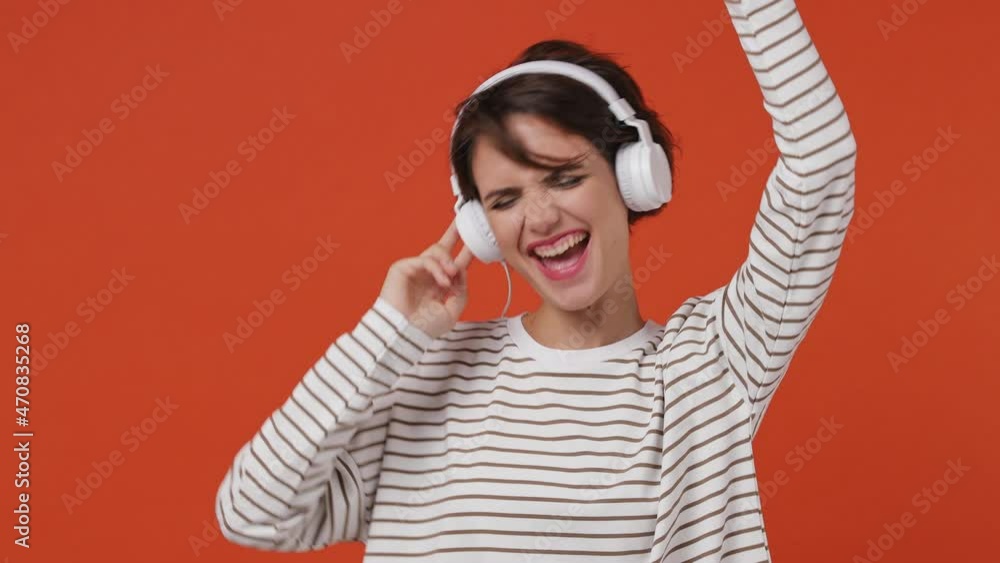 Vivid cheerful young brunette woman 20s years old wears striped shirt listen music in headphones dance sing song fooling around gesticulating hands isolated on plain orange background studio portrait