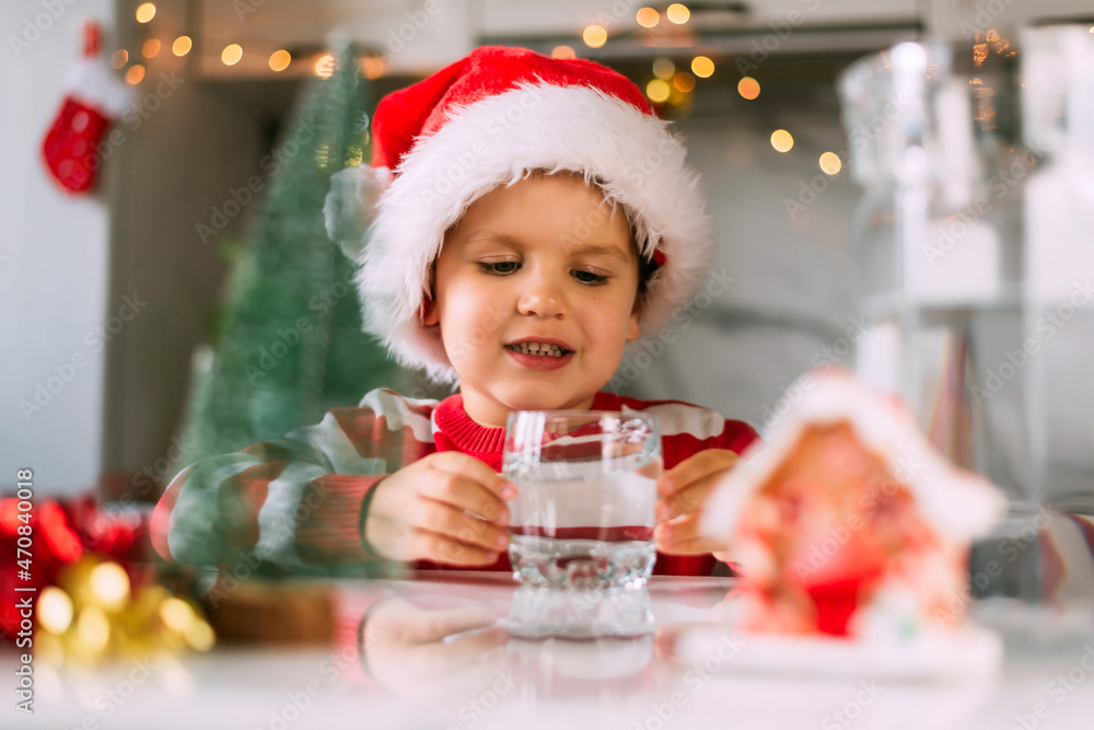 Happy funny toddler boy in a red Santa hat drinking filtered water from a glass in the kitchen. Holidays, health concept.