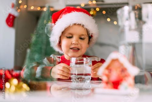 Happy funny toddler boy in a red Santa hat drinking filtered water from a glass in the kitchen. Holidays, health concept.