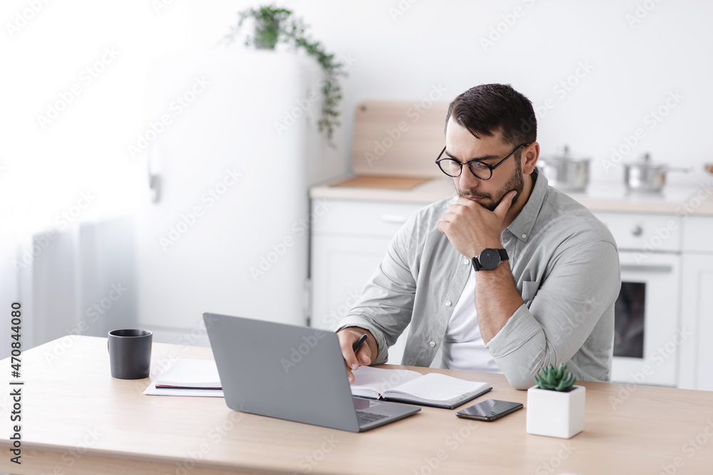 Busy pensive middle aged european businessman in glasses working at computer on minimalist kitchen interior - obrazy, fototapety, plakaty 