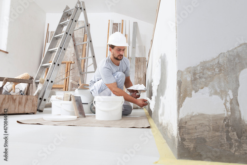 man plasterer construction worker at work, takes plaster from bucket and puts it on trowel to plastering the wall, wears helmet inside the building site of a house photo