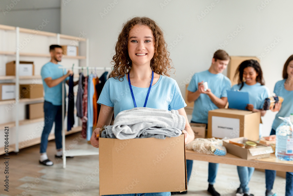 Volunteering concept. Happy young woman in uniform holding cardboard box with clothes for donation Volunteering concept. Happy young woman in uniform holding cardboard box with clothes for donation