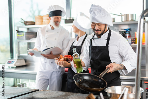 Smiling asian chef holding frying pan and olive oil near cooktop and blurred colleagues in kitchen