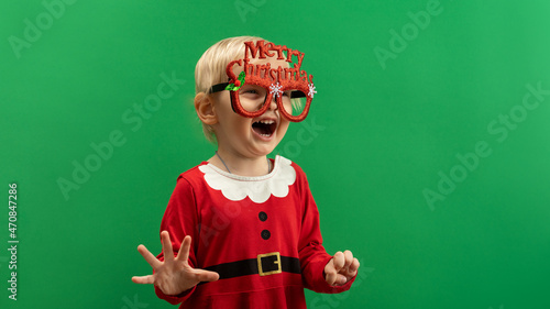  Happy Toddler girl in red Christmas costume and glasses laughs on green background COPYSPACE