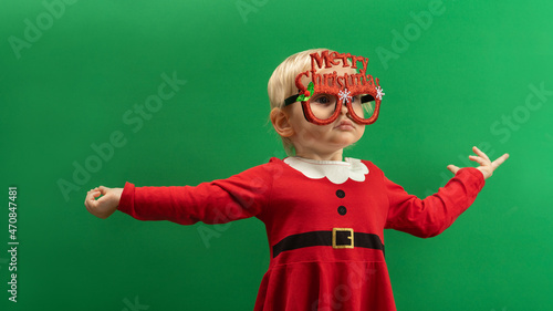  Happy Toddler girl in red Christmas costume and glasses smiling on green background COPYSPACE